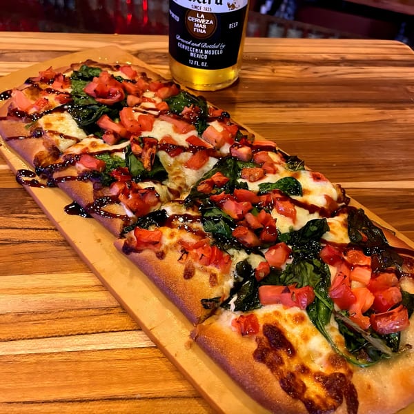 Flatbread pizza with spinach, diced tomatoes, melted cheese, and balsamic glaze, next to a beer bottle.
