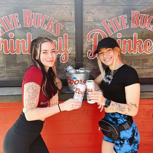Two smiling women holding Coors Light cans in front of the FIVE BUCKS DRINKERY sign.