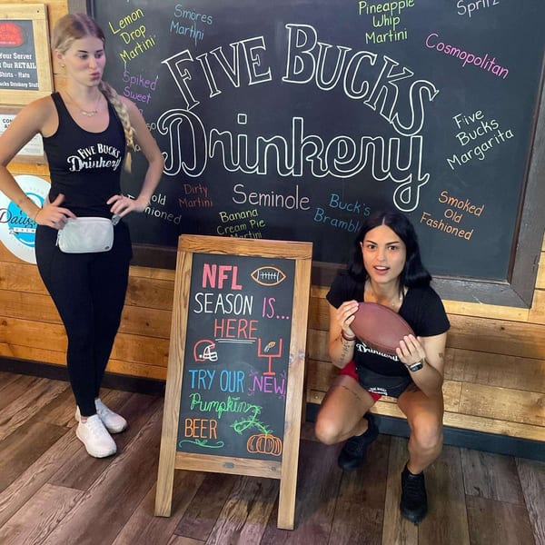 Two women pose with a football and a chalkboard sign advertising NFL season and pumpkin beer at FIVE BUCKS DRINKERY.