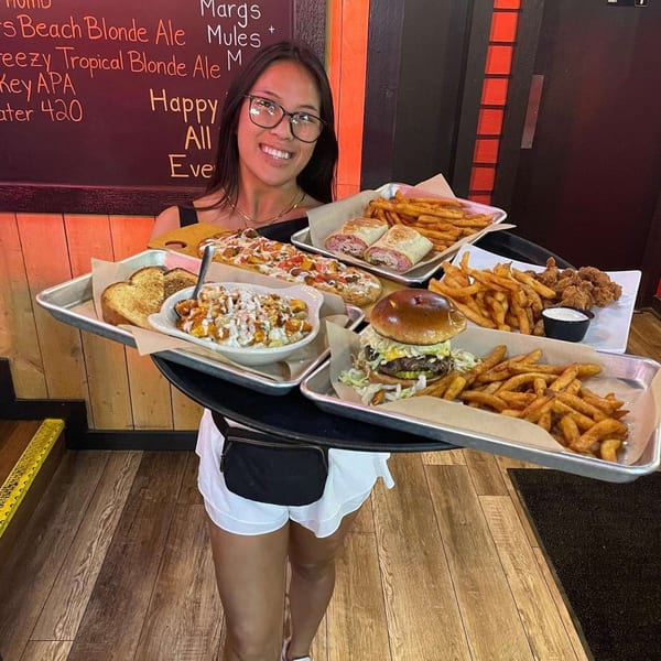 Smiling woman carrying a large tray with multiple bar-and-grill dishes, including burgers, fries, and wraps.