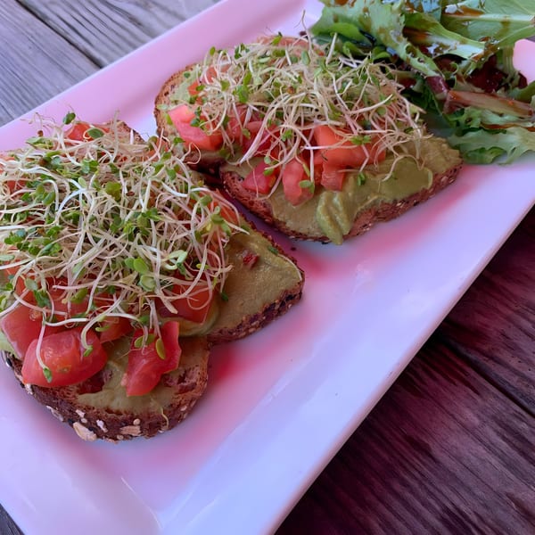 Avocado toast with diced tomatoes and sprouts, served with a side salad on a white plate.