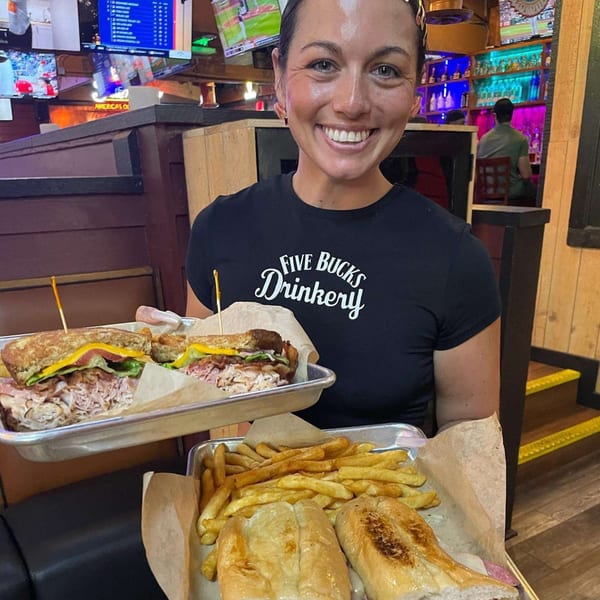 A smiling FIVE BUCKS DRINKERY server holds two trays of sandwiches and fries in a lively bar.