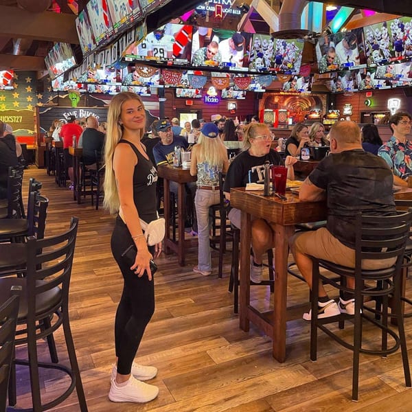 A woman smiles in a lively bar-and-grill with many TVs showing sports and patrons at high-top tables.