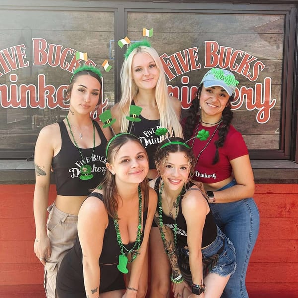 Five women wearing St. Patrick's Day themed accessories pose in front of the FIVE BUCKS DRINKERY sign.
