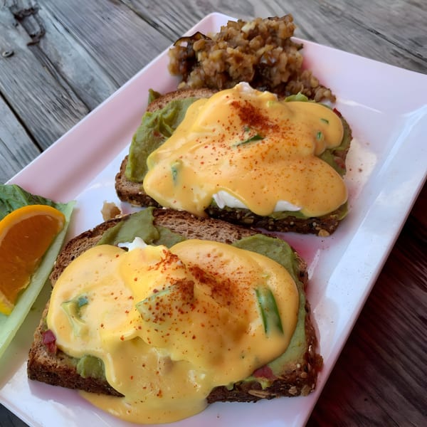 Avocado toast with poached eggs and hollandaise sauce, served with hash browns and an orange slice.
