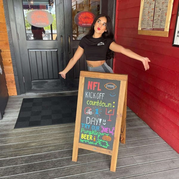 A woman in a FIVE BUCKS DRINKERY shirt poses next to an "NFL Kick Off" sign.